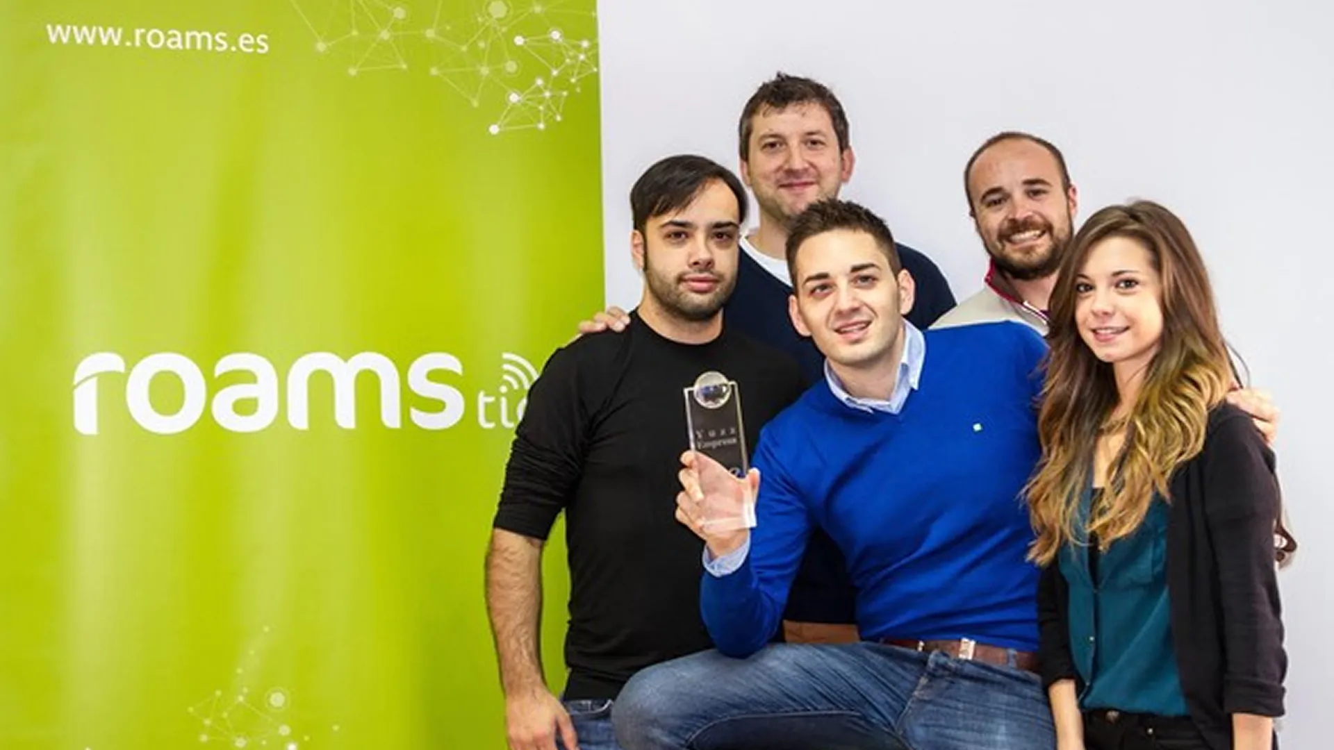 Equipo de Roams posa ante un roll-up verde de la empresa; uno de ellos sostiene un premio.