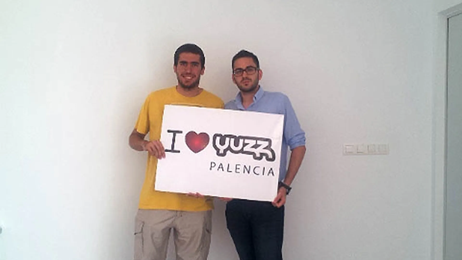 Eduardo Delgado y Alberto Martín sostienen un cartel con el lema I love Yuzz Palencia en una sala blanca.
