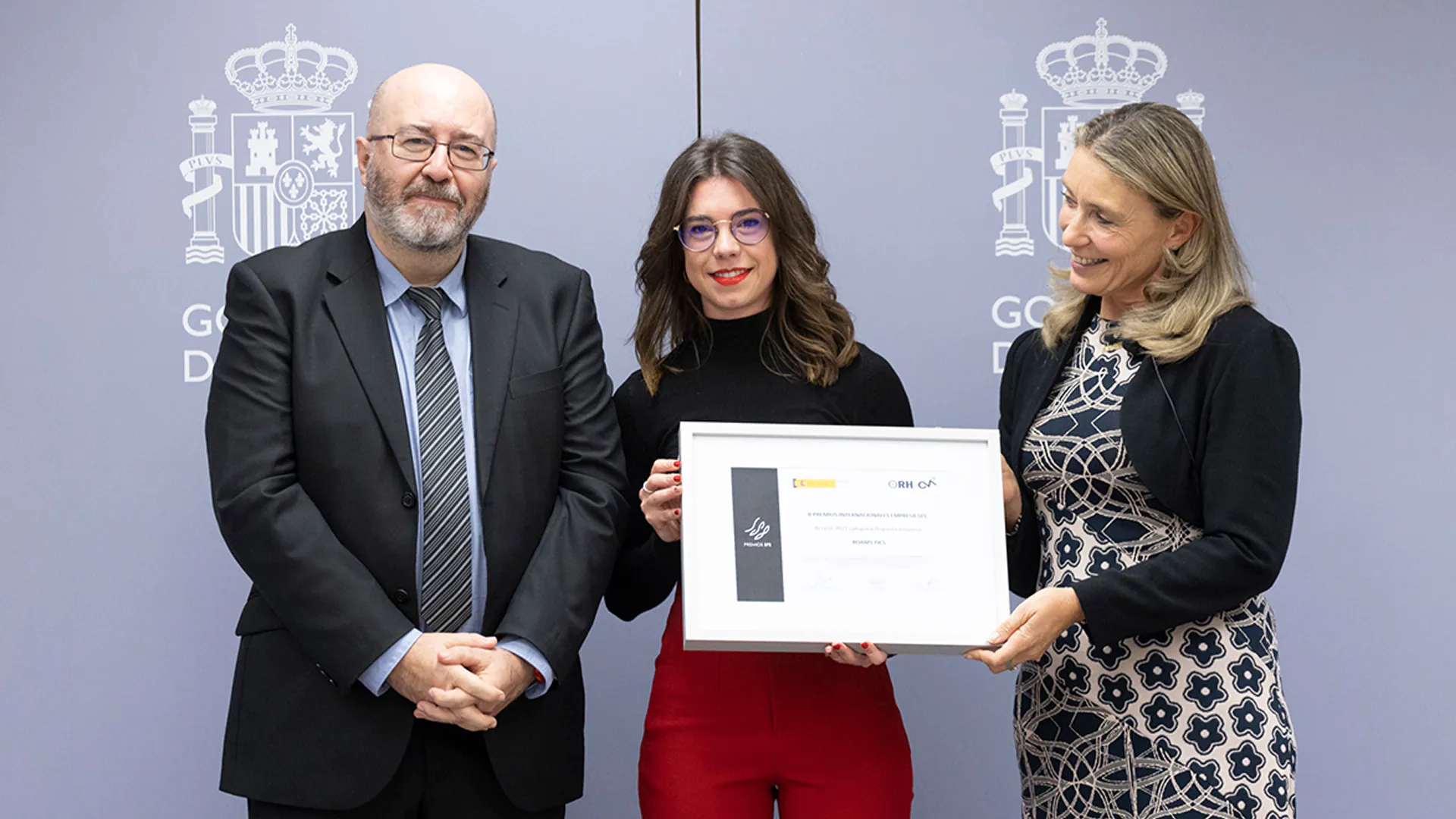 Esther Gómez recibe un diploma enmarcado junto a un hombre y otra mujer ante un fondo del Gobierno de España.