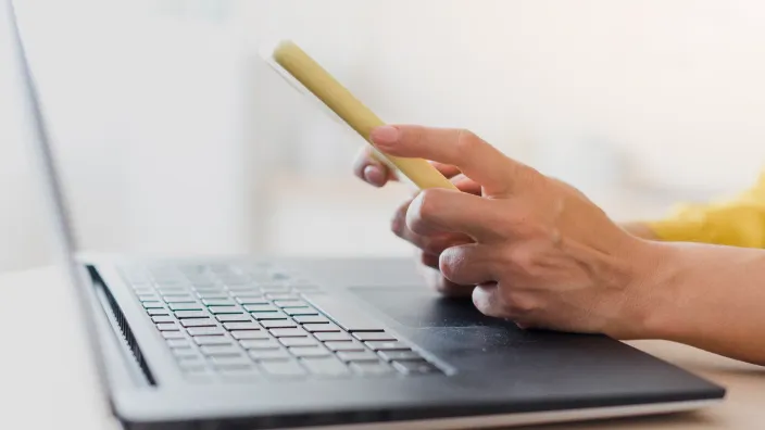 Mano de una persona usando un teléfono móvil frente a un ordenador portátil, representando el uso conjunto de internet y telefonía.