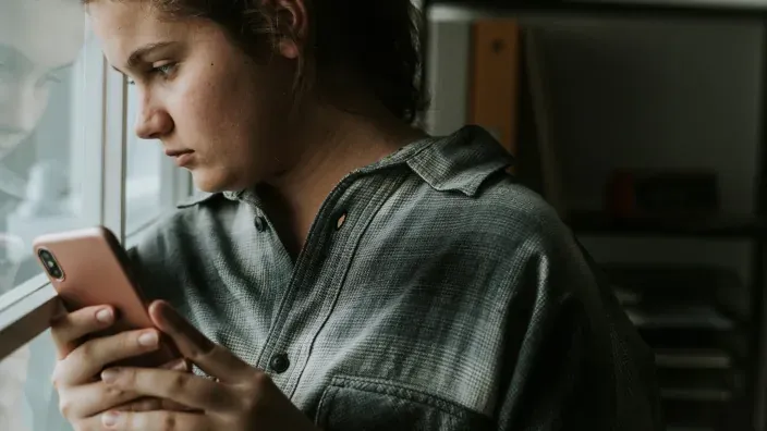 Adolescente triste mirando su móvil junto a una ventana, víctima de ciberbullying o acoso a través de las redes sociales.