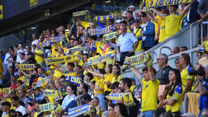 Aficionados del Cádiz CF animando a su equipo en la grada con camisetas y bufandas amarillas.