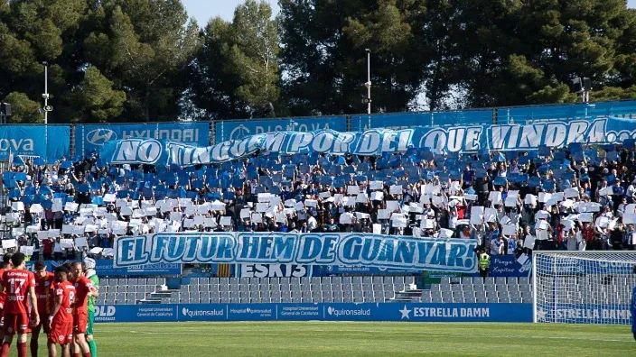 Afición del CE Europa animando en la grada con pancartas azules y blancas