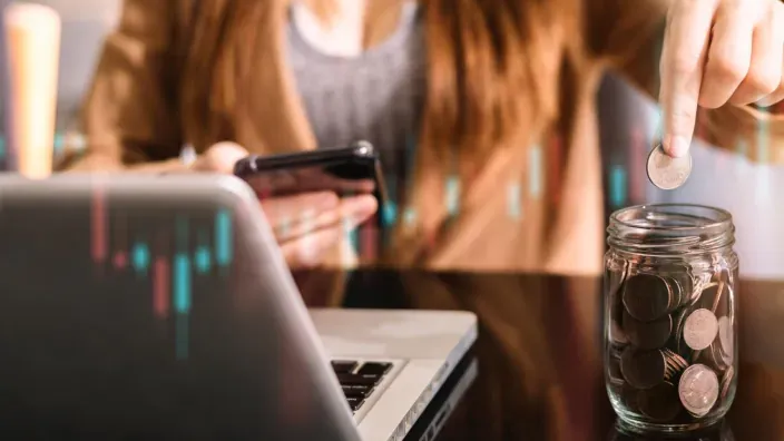 Persona guardando monedas en un tarro mientras usa el móvil junto a un ordenador portátil, representando el ahorro y la gestión de finanzas personales.