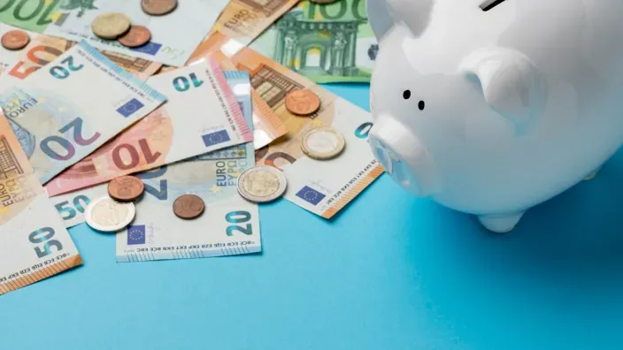 Hucha cerdito blanca con dinero en efectivo en billetes y monedas sobre fondo azul.