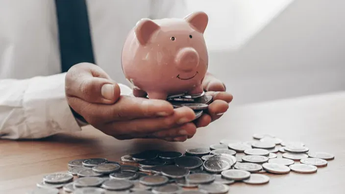 Manos sosteniendo una hucha con forma de cerdito sobre una mesa llena de monedas, símbolo de ahorro y finanzas personales.