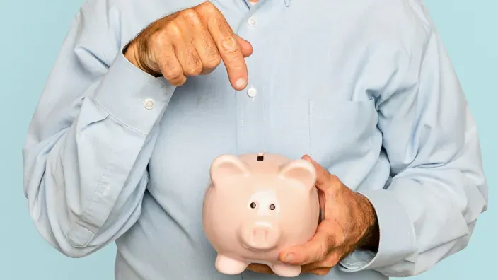 Hombre señalando una hucha de cerdito rosa para representar la importancia del ahorro y las finanzas personales.