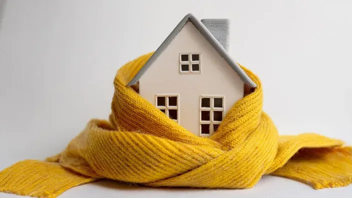 Maqueta de casa envuelta en bufanda de lana amarilla simbolizando el aislamiento térmico y el calor.