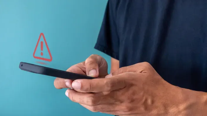 Persona con smartphone e icono de alerta roja, representando un aviso de seguridad o estafa digital.