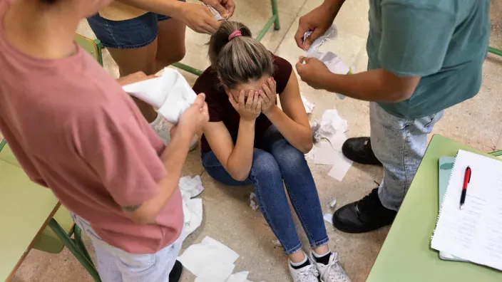 Una chica llorando en el suelo de clase mientras otros estudiantes la rodean y le tiran papeles, una escena de acoso escolar.