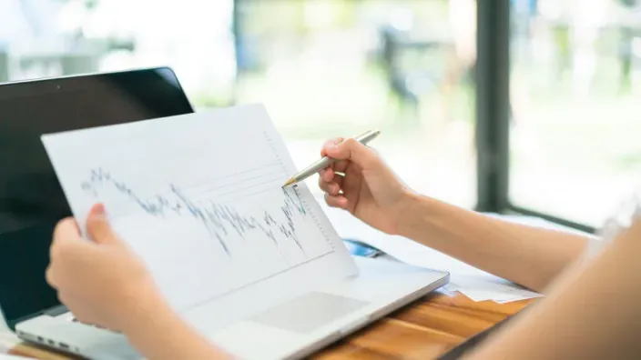 Persona revisando un gráfico de evolución de mercados financieros junto a un ordenador portátil.