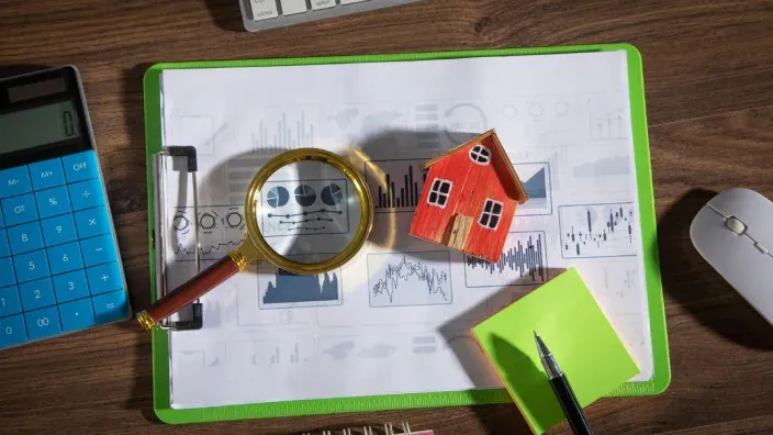 Lupa sobre gráficos financieros y maqueta de casa roja en escritorio con calculadora.