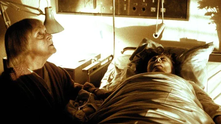 Una mujer mayor sostiene la mano de otra enferma en una escena inquietante iluminada con luz amarilla de la serie El reino.