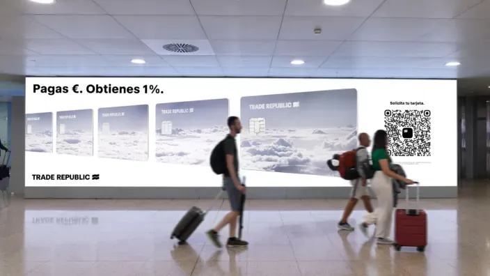Publicidad de Trade Republic en un aeropuerto con el eslogan “Pagas €. Obtienes 1%” y personas caminando con maletas frente al anuncio.