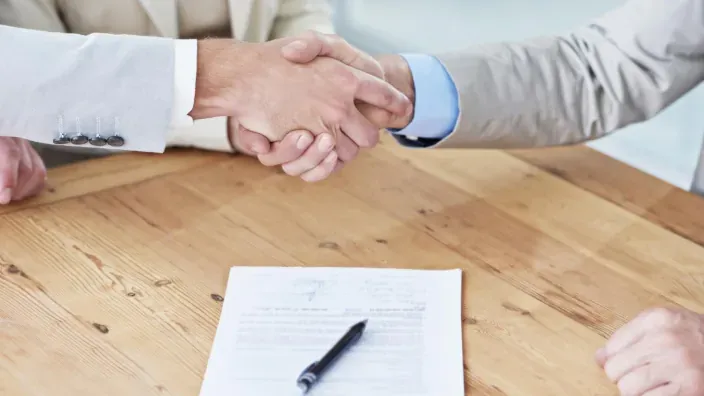 Dos personas estrechándose la mano sobre una mesa con un contrato y un bolígrafo