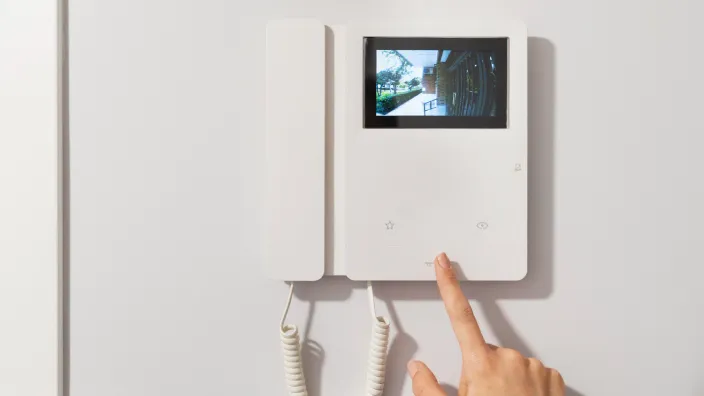 Mano pulsando botón de videoportero con pantalla mostrando entrada exterior de una vivienda.