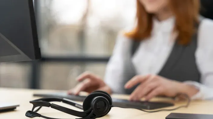 Auriculares con micrófono sobre escritorio de oficina y mujer trabajando al fondo desenfocada.