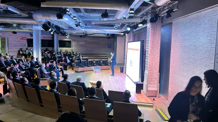Ponente realizando una presentación en un auditorio moderno, con pantallas grandes y público siguiendo la exposición.