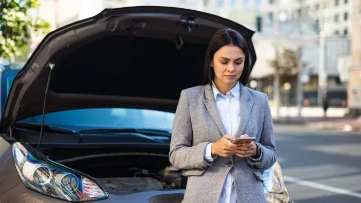 Mujer consultando el móvil frente a un coche averiado con el capó abierto en la calle.