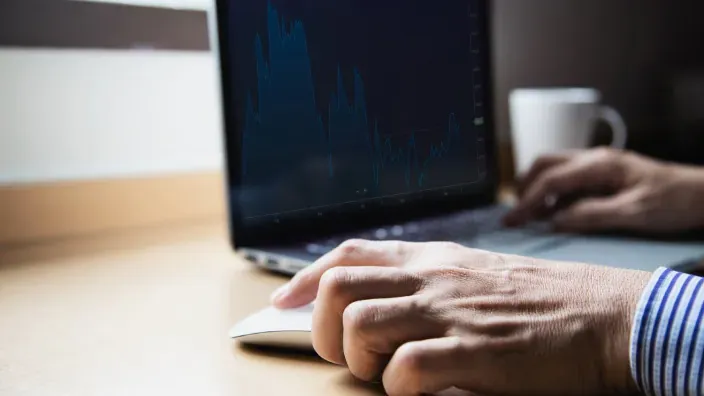 Primer plano de trader operando en bolsa con gráficos financieros en la pantalla del portátil.