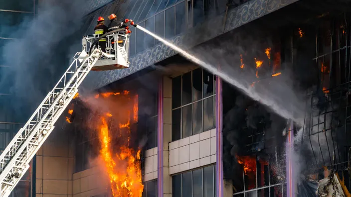 Bomberos trabajando desde una escalera hidráulica para apagar un incendio en un edificio en llamas.