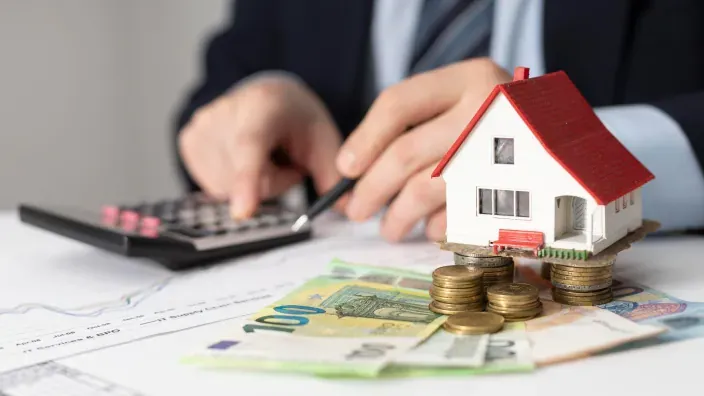 Persona calculando los gastos de una hipoteca o el precio de una vivienda con una calculadora, dinero y una casa en miniatura.