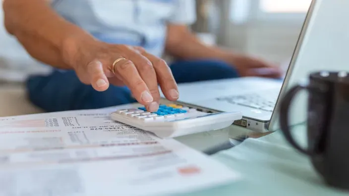 Hombre mayor calculando gastos domésticos con calculadora, facturas y portátil en casa.