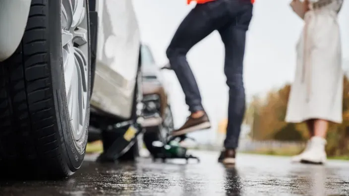 Pareja intentando cambiar rueda pinchada de coche averiado en el arcén de carretera mojada.