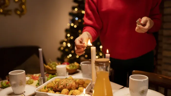 Mujer encendiendo velas en mesa preparada para cena de Navidad con comida y árbol de fondo.