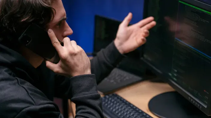 Ciberdelincuente hablando por teléfono móvil mientras ejecuta código malicioso en el ordenador.