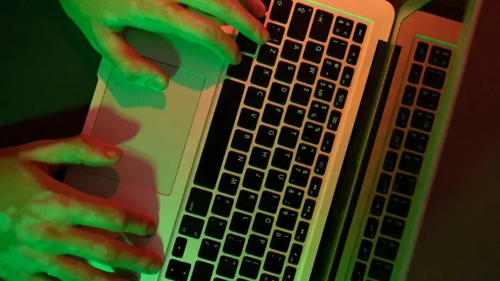 Hacker utilizando un ordenador portátil bajo luces verdes y rojas, representando un ciberataque o hacking.