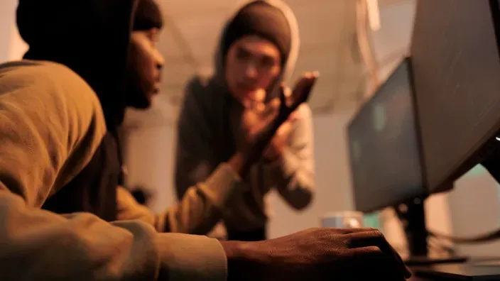 Dos ciberdelincuentes conversando frente a varios ordenadores mientras planifican una posible estafa digital.