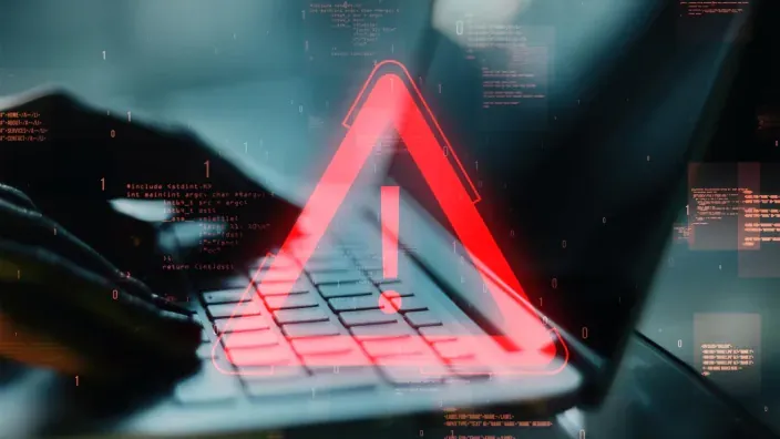 Alerta de seguridad roja con signo de exclamación sobre portátil hackeado con código binario de fondo.