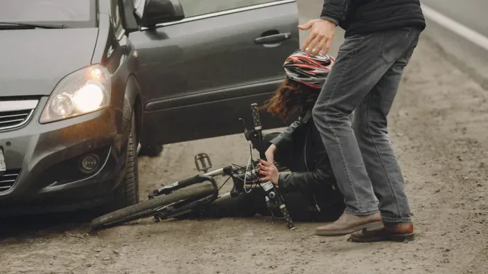 Ciclista caído en la calzada junto a un coche y otra persona