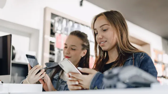 Dos chicas jóvenes en una tienda de telefonía comparando las características de varios modelos de smartphones antes de comprar.