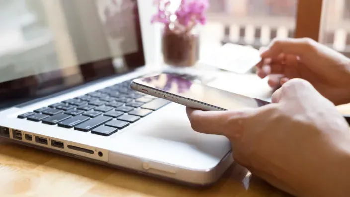 Persona realizando una compra online con el móvil frente a un ordenador portátil.