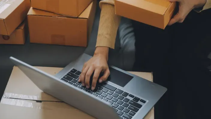 Persona gestionando pedidos online con un portátil rodeado de cajas de envíos.