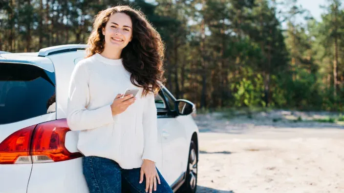 Mujer joven sonriente apoyada en coche blanco en el bosque usando el móvil durante un viaje.