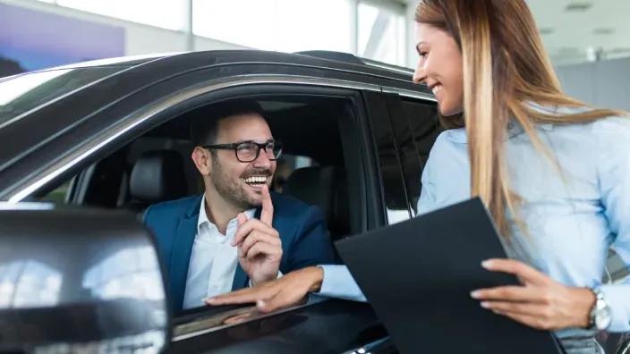 Hombre sonriente al volante de coche nuevo hablando con vendedora en concesionario.