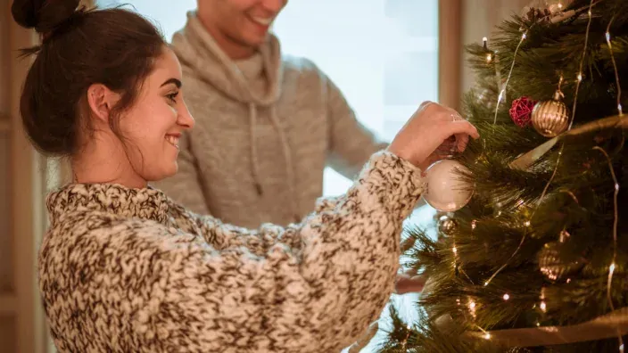 Pareja joven sonriente decorando juntos el árbol de Navidad con bolas plateadas y luces.