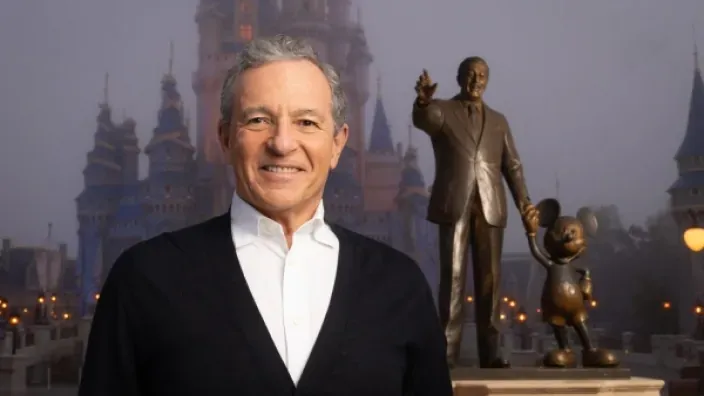 Bob Iger CEO de Disney posando sonriente frente al castillo y estatua de Walt Disney y Mickey Mouse.