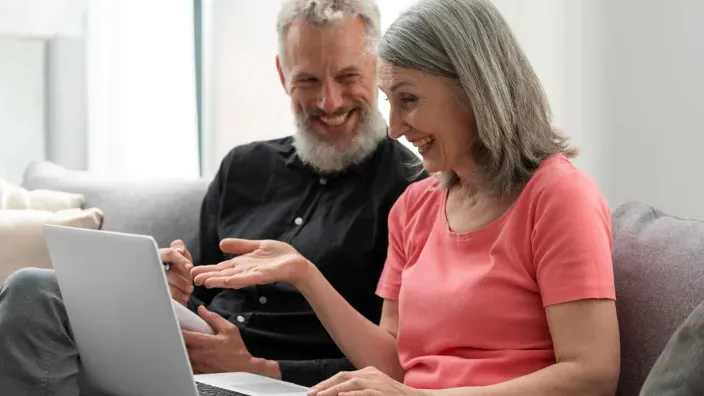 Pareja de jubilados sonríe mientras revisa información sobre en su ordenador portátil.