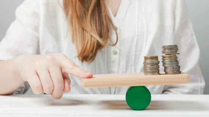 Persona equilibrando pilas de monedas sobre una tabla para representar el balance financiero y el ahorro personal.