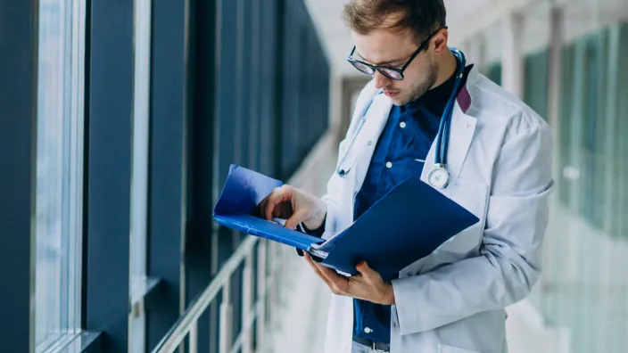 Médico con bata blanca y estetoscopio revisa documentos médicos en carpeta azul en pasillo acristalado del hospital.