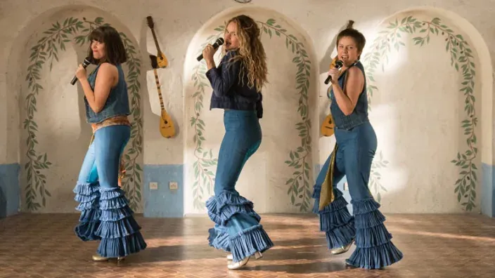 Tres mujeres cantando y bailando con micrófonos en una escena musical ambientada en un interior decorado con motivos florales.