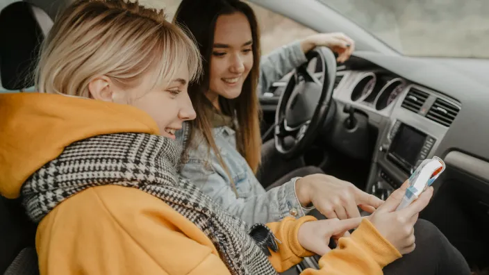 Dos amigas consultando el móvil dentro de un coche, probablemente usando el GPS o buscando información para su viaje.