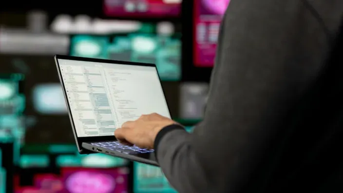 Hacker trabajando en un portátil frente a múltiples monitores de un centro de datos.