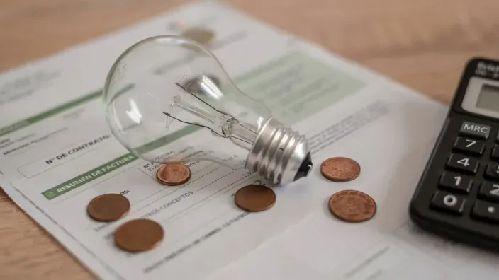 Bombilla sobre una factura de electricidad junto a monedas y una calculadora, simbolizando el coste de la luz