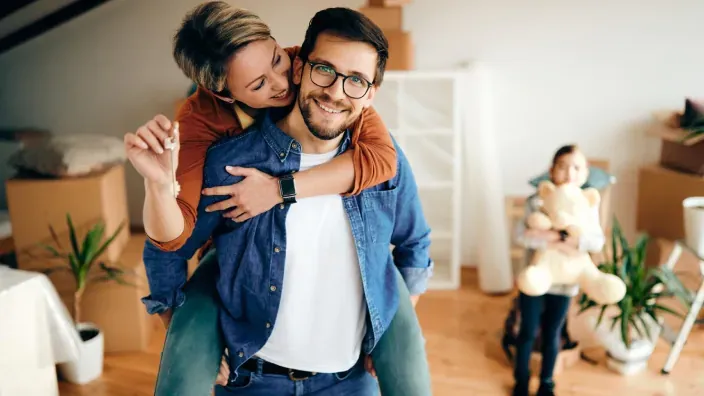 Pareja feliz en su nuevo hogar durante la mudanza con cajas y una niña al fondo.