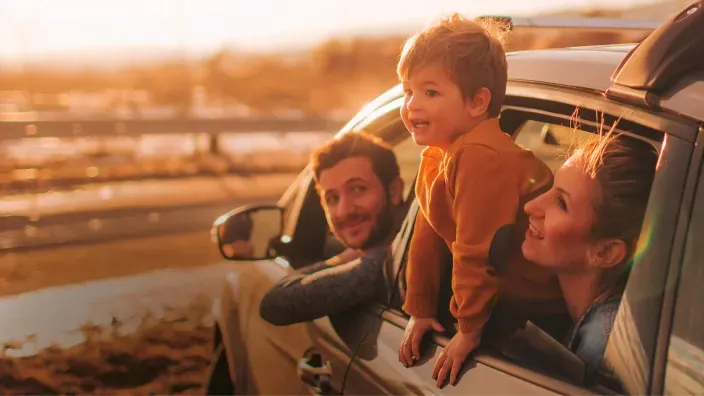 Familia feliz asomada por las ventanillas de su coche al atardecer para representar los viajes y seguros de auto.
